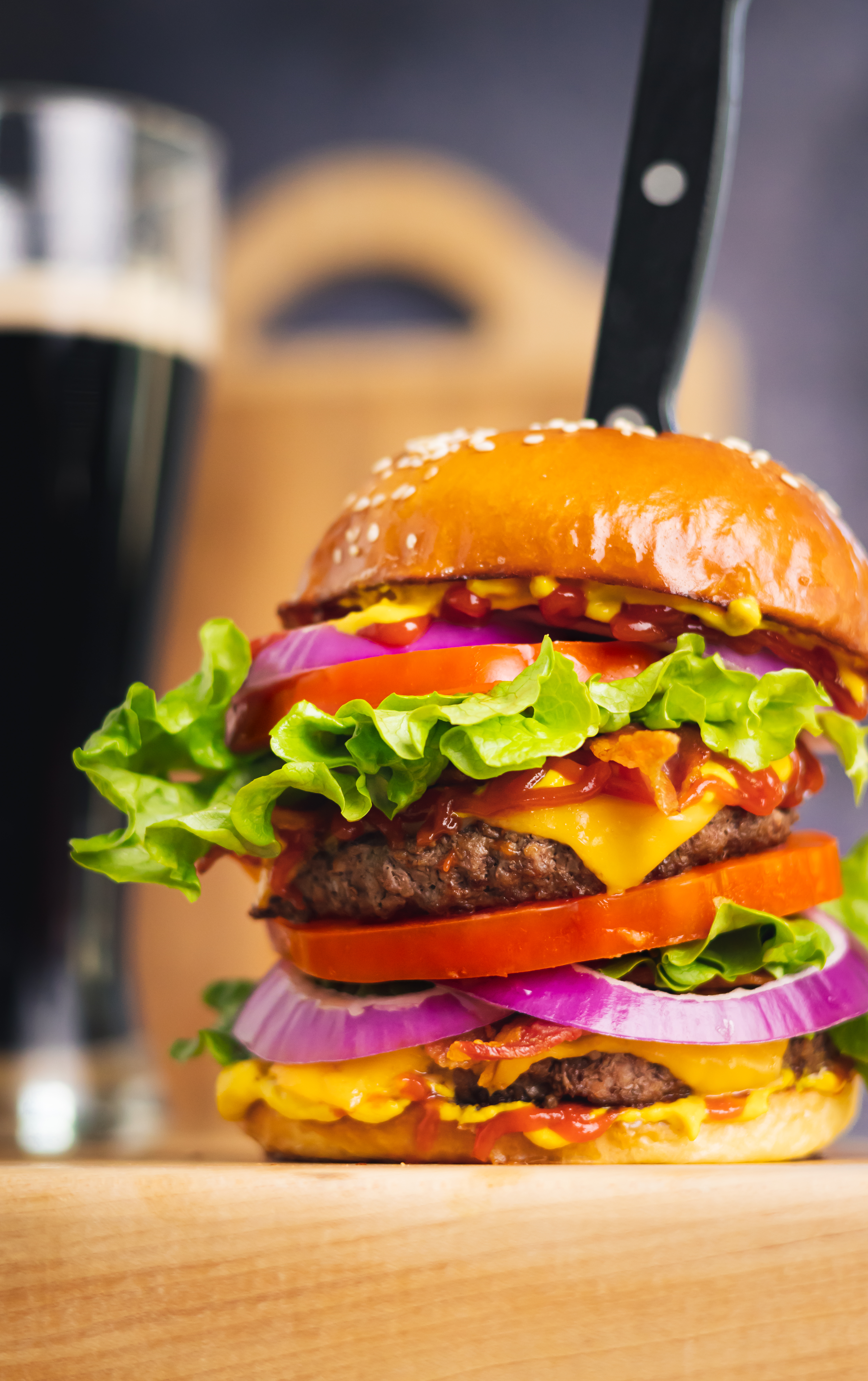 a placeholder image of a steak knife placed through the top of a large cheeseburger with many toppings and a blurry beer in a glass behind it